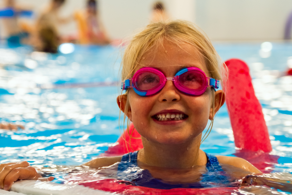 A young girl smiles at the camera while swimming with a woggle