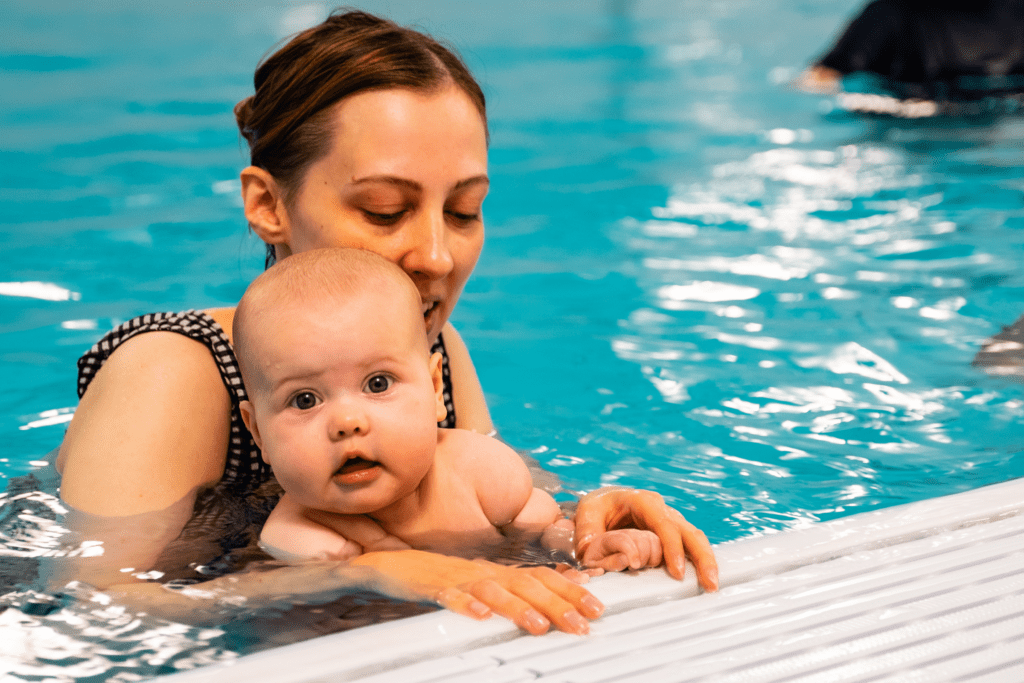 A mum helps a baby to hold on to the side of the pool during a baby swimming lesson