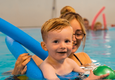 A young boy swims with the support of his mum and a woggle while holding a green ball