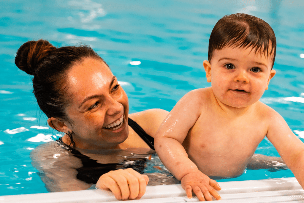 A toddler and mum hold on to the side of the swimming pool while the young boy smiles at the camera