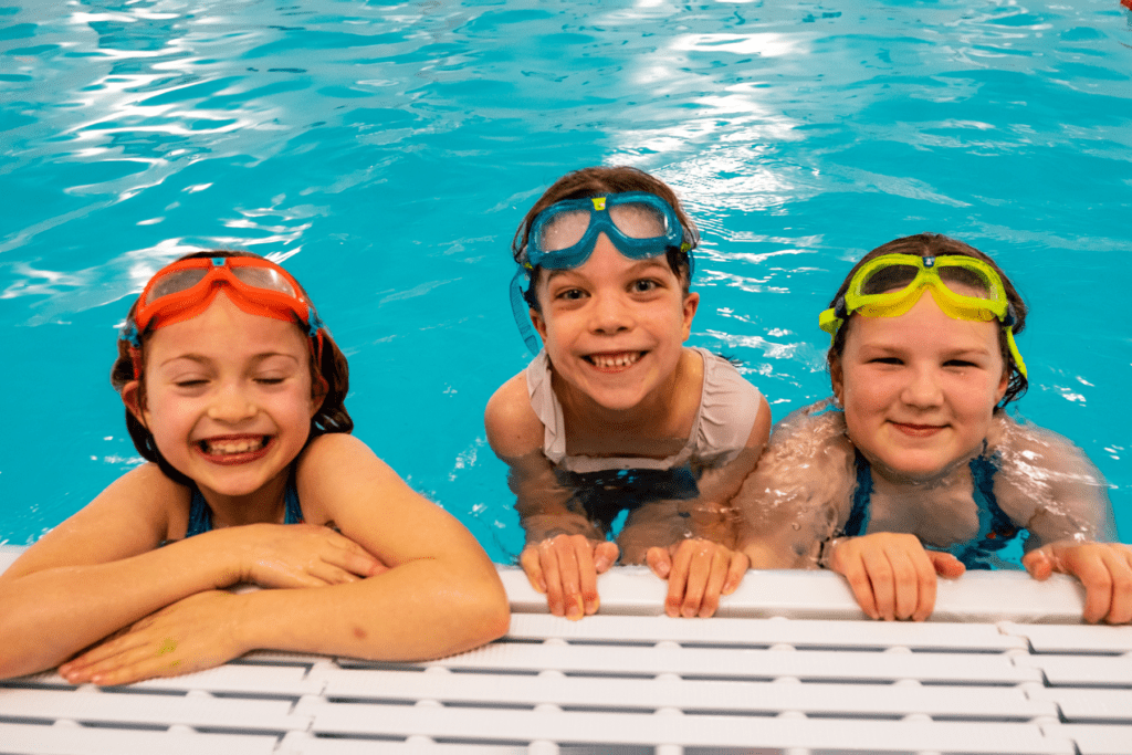Three girls in a swimming pool smile at the camera while holding on the the side of the pool