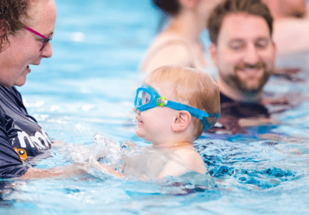 A Nemo Swimming instructor praises a young boy during his swimming lesson while a man smiles in the background