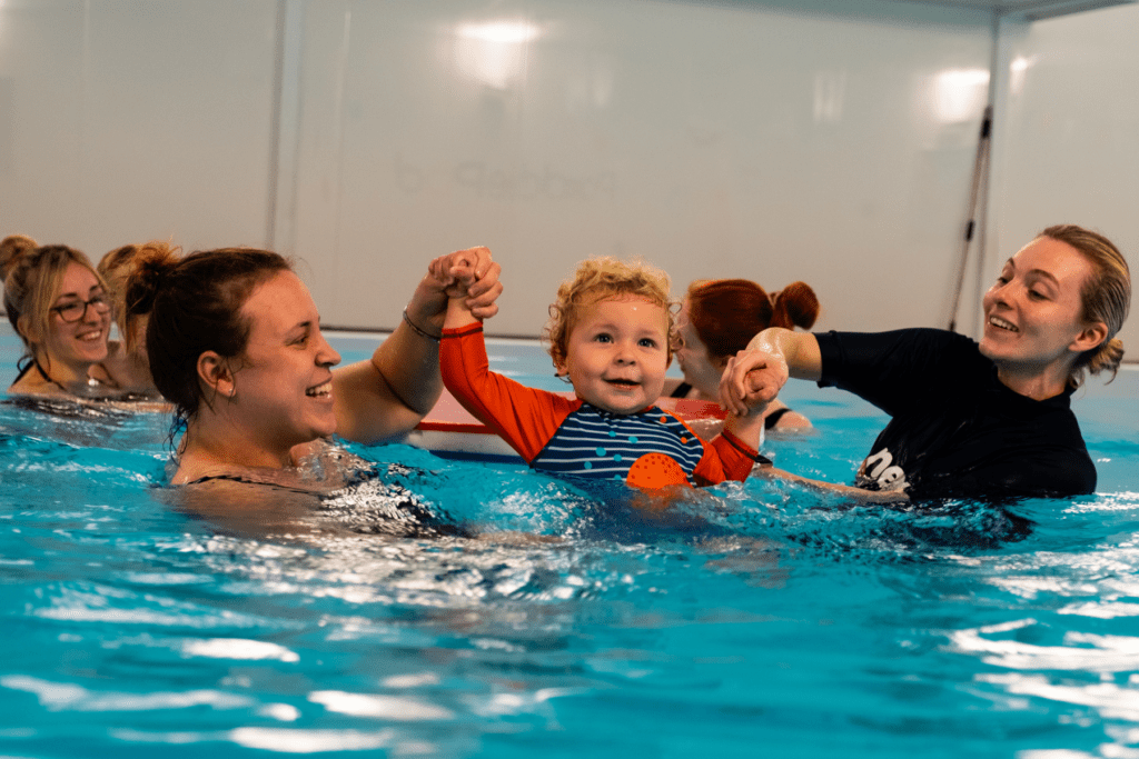 A smiley toddler jumps into the swimming pool with the support of his mum and swimming teacher