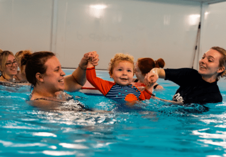 A smiley toddler jumps into the swimming pool with the support of his mum and swimming teacher