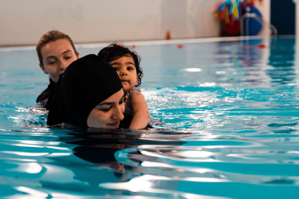 A woman swims with a toddler on their back while an instructor supports
