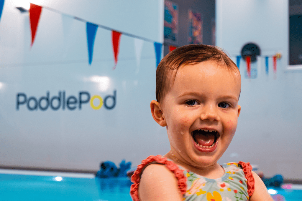 A toddler smiles at the camera while enjoying a swimming lesson