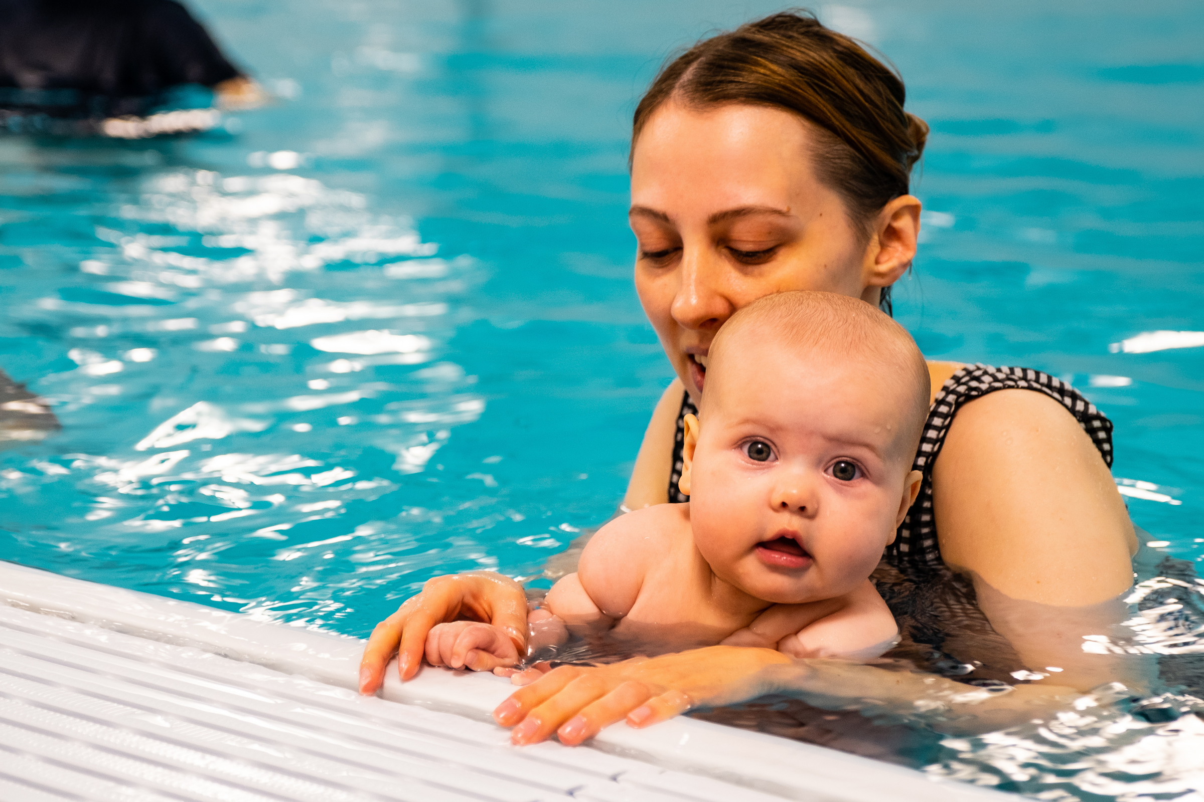 A mum helps a baby to hold on to the side of the pool during a baby swimming lesson