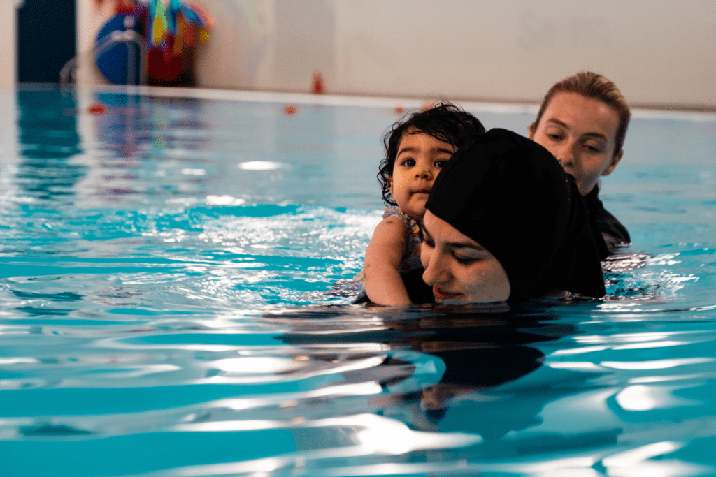 A woman swims with a toddler on their back while an instructor supports