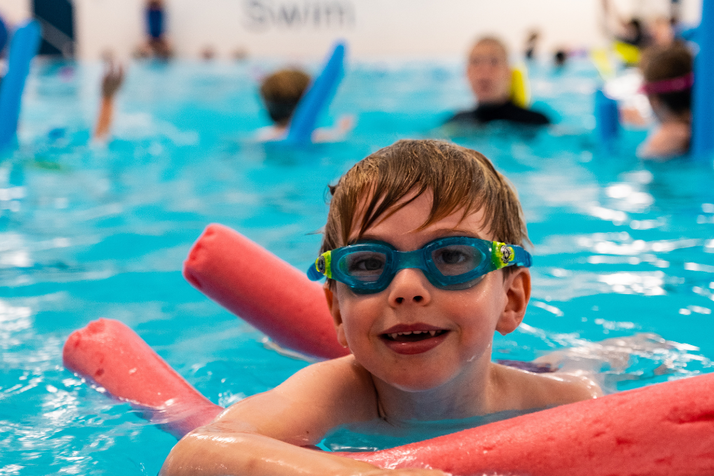 A pre-school swimmer with blue goggles and a red woggle smiles at the camera