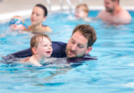 A dad supports his toddler as they move through the water during a swimming class