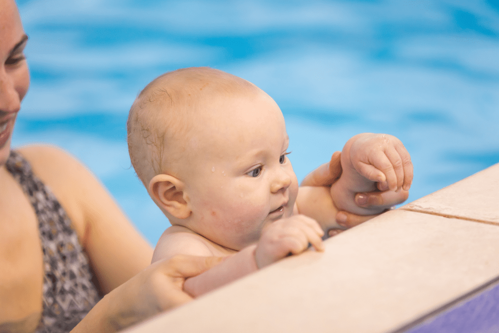 A baby holds on to the side of the swimming pool while mum supports
