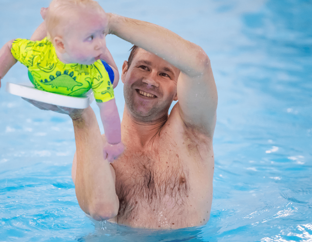 A dad supports baby on a float and lifts him out of the swimming pool in a surfing action