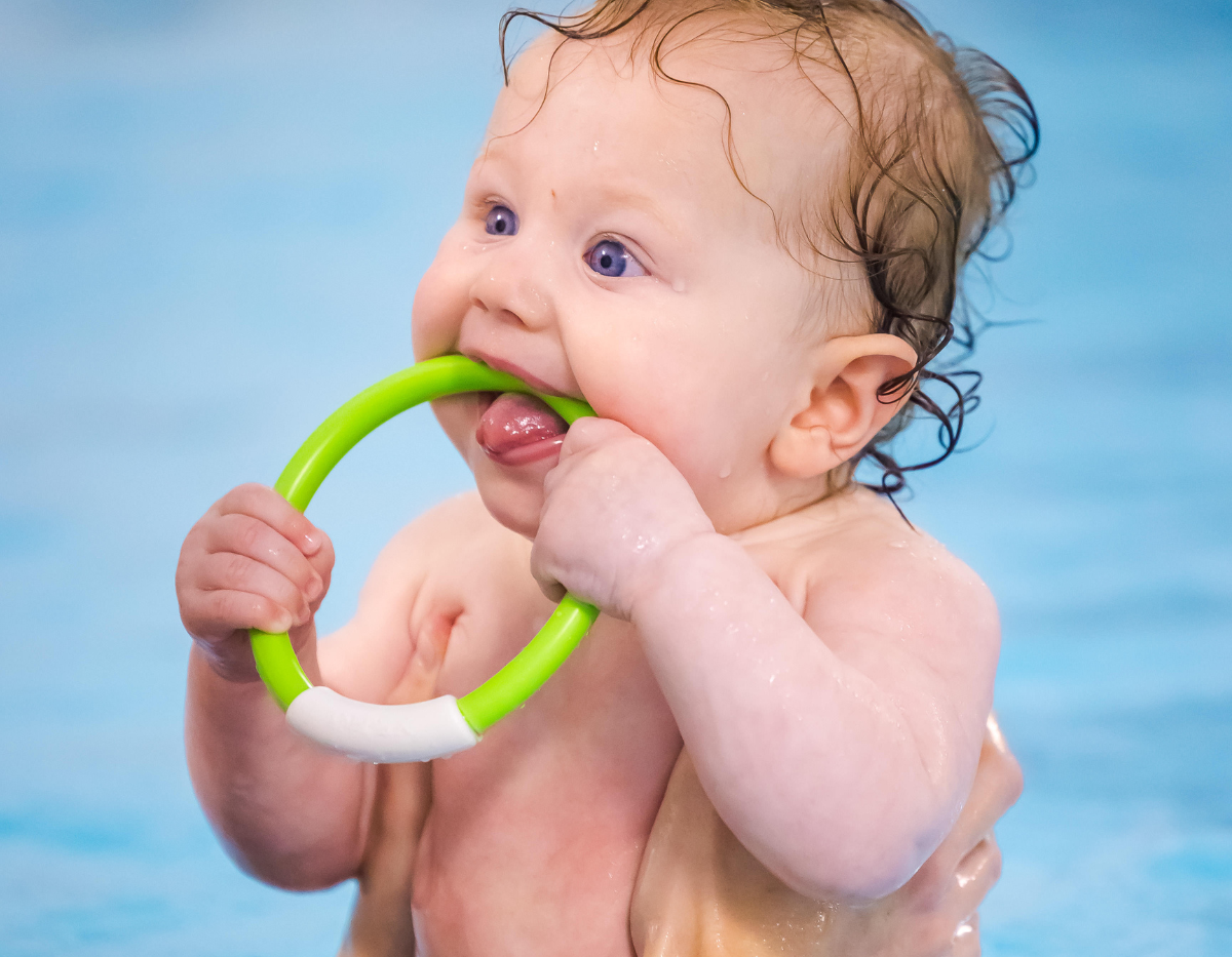 A young baby chews on a green ring toy while in the swimming pool