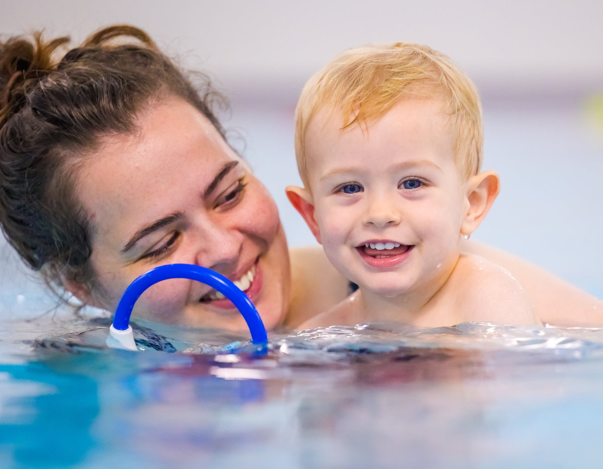 A toddler and mum hold a blue ring and smile during a swimming class