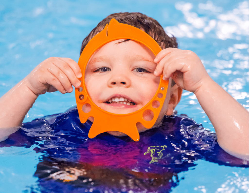 A young boy in a swimming pool looks through a hoop and smiles at the camera