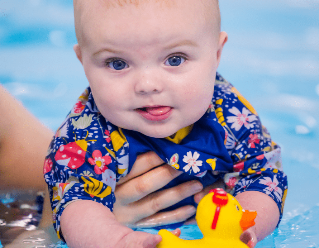 A baby holds a rubber duck and smiles at the camera while in the swimming pool