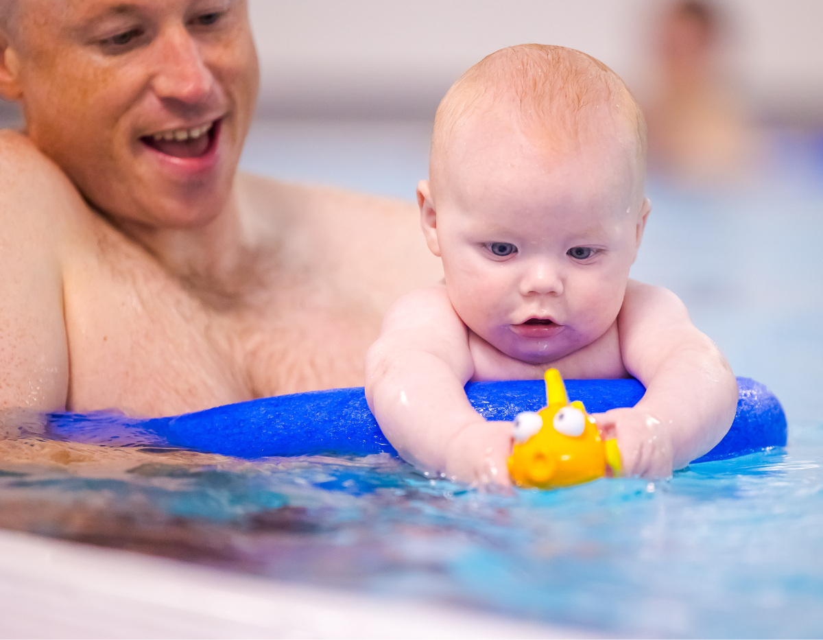A baby reaches out for a toy while being supported by dad in the swimming pool