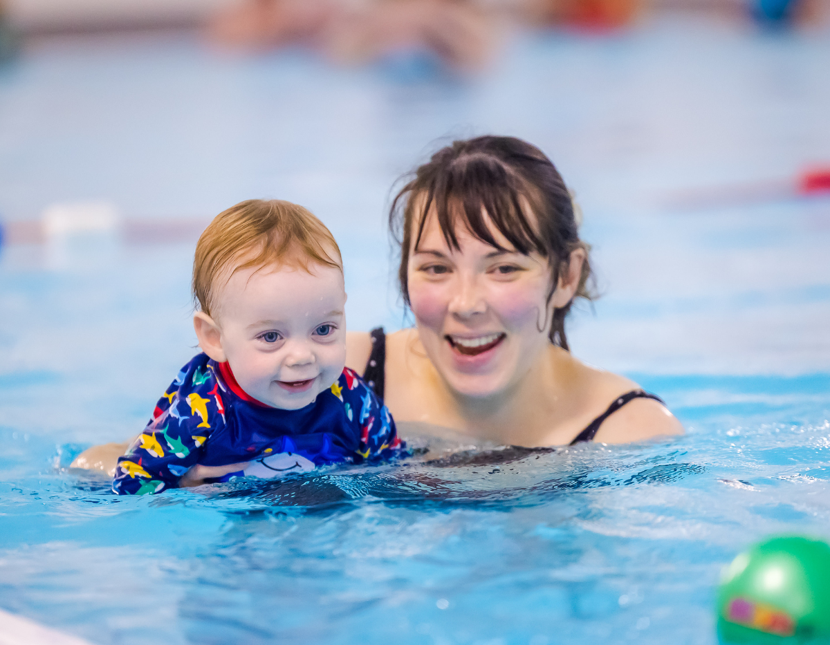 A baby and mum smile and follow a green ball in the swimming pool