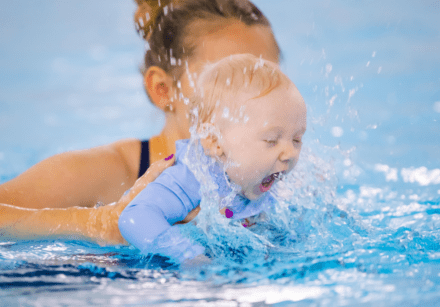 A baby splashes and smiles in the swimming pool with mum