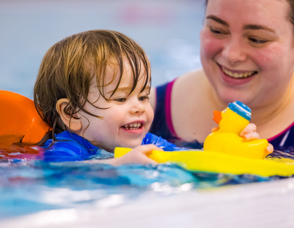 A toddler smiles in the swimming pool while using a float to transport a rubber duck