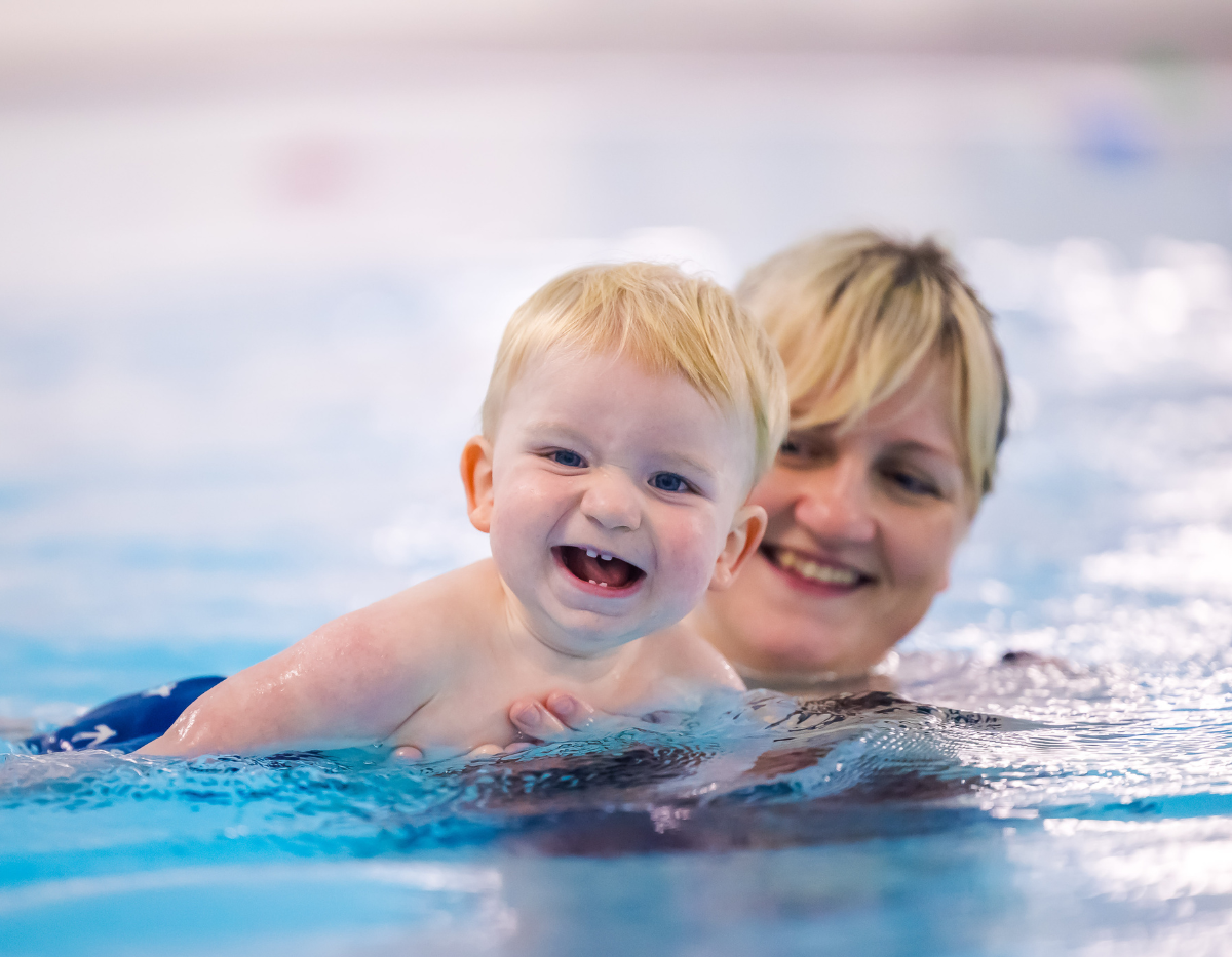 A toddler and mum move through the swimming pool while smiling at the camera