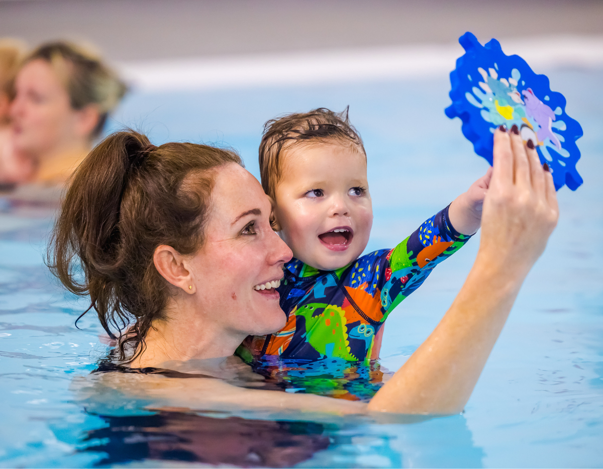 A mum supports a toddler in the swimming pool while they hold a mirror and smile into it