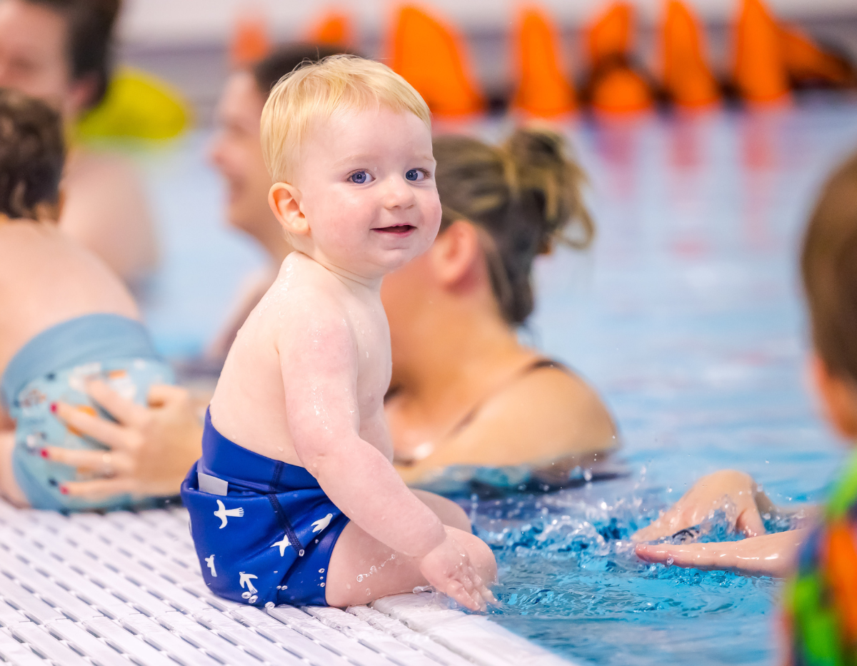 A toddler in blue shorts sits on the side of the swimming pool and smiles at the camera