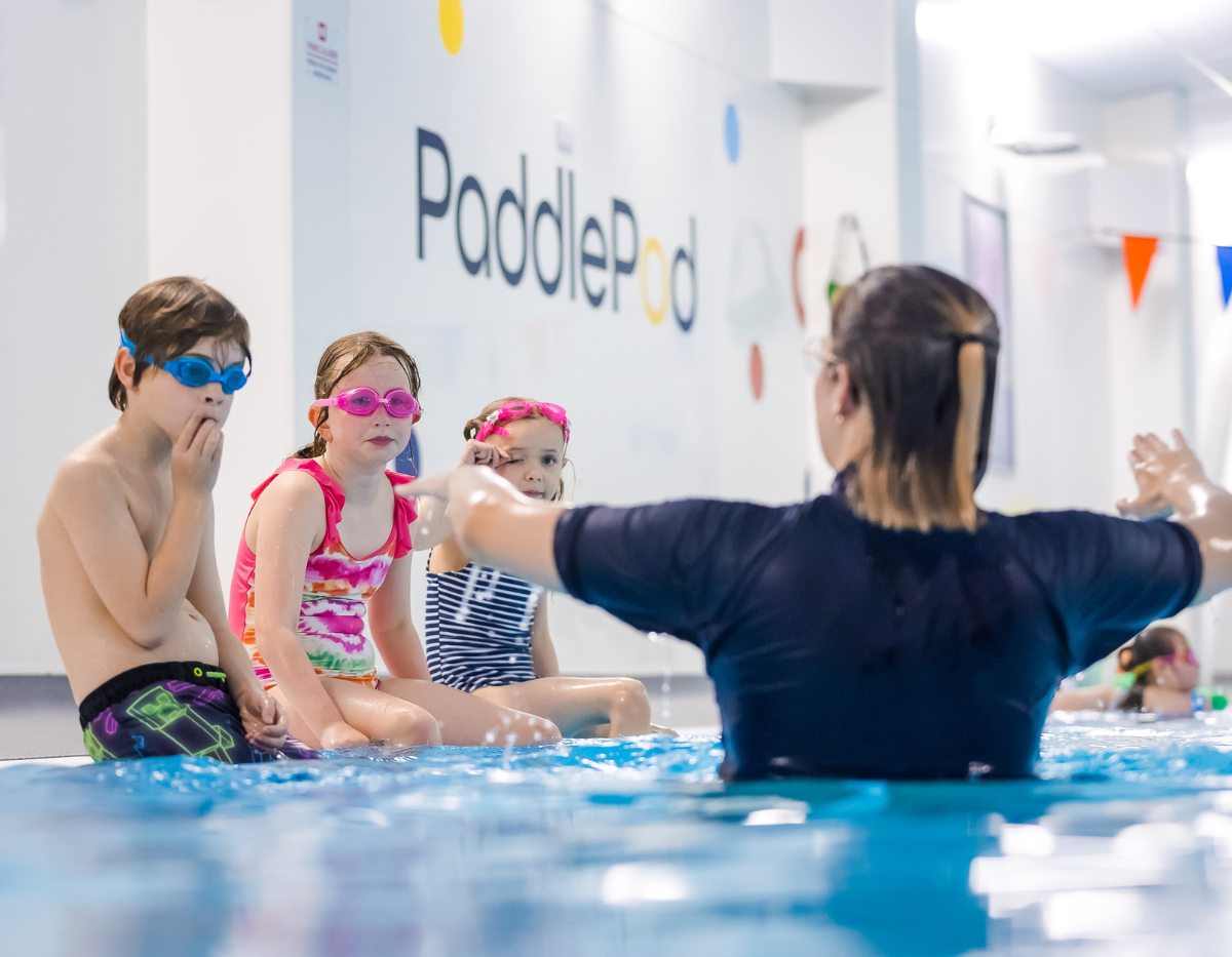 Three children sit on the side of the pool while their swimming teacher demonstrates