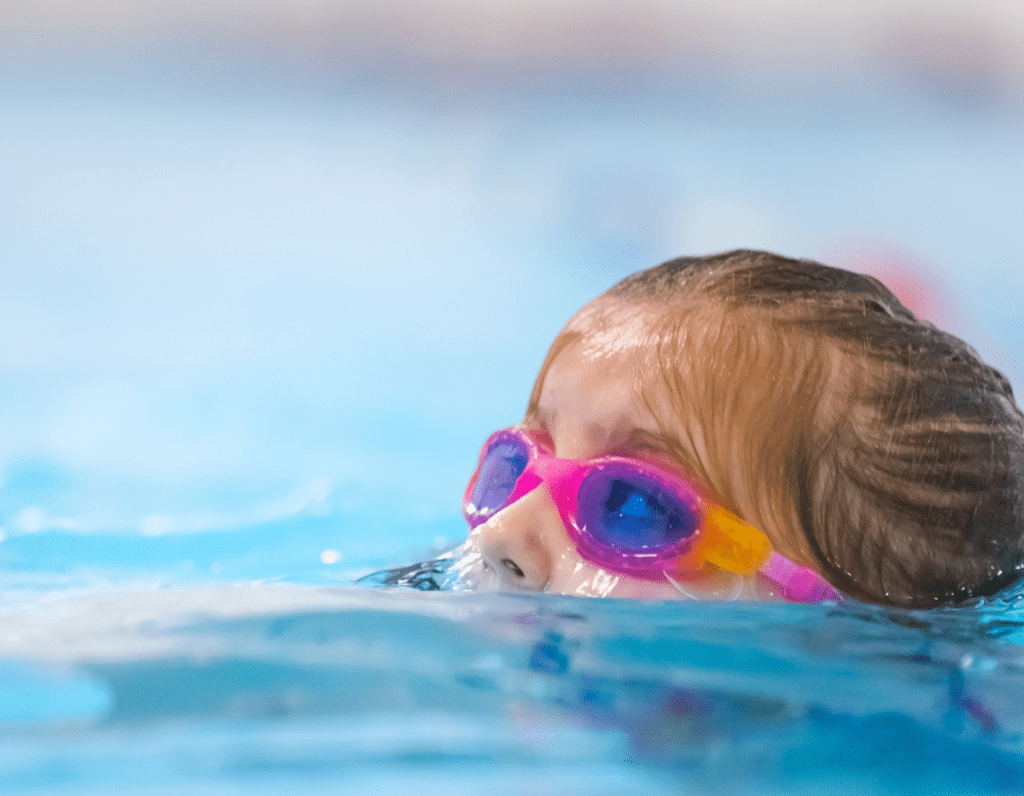 A girl with pink goggles emerges from the pool to take a breath