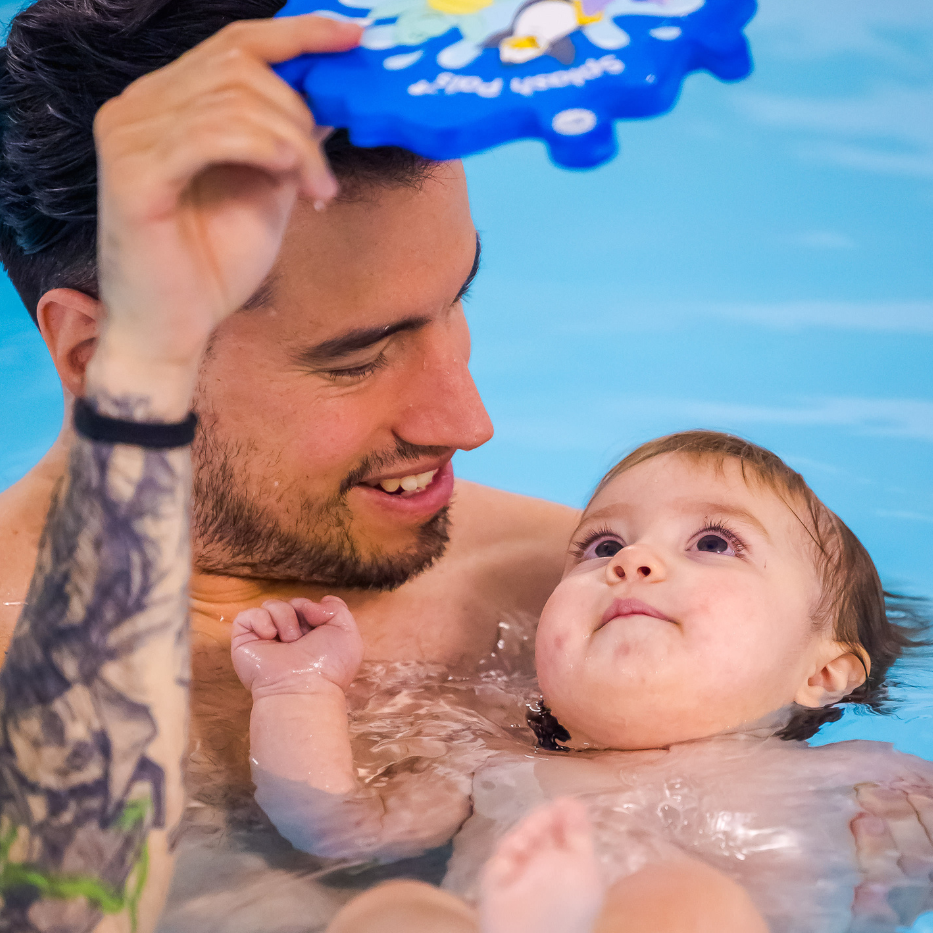 A baby floats on her back in a swimming pool while being supported by dad and looking at herself in a mirror