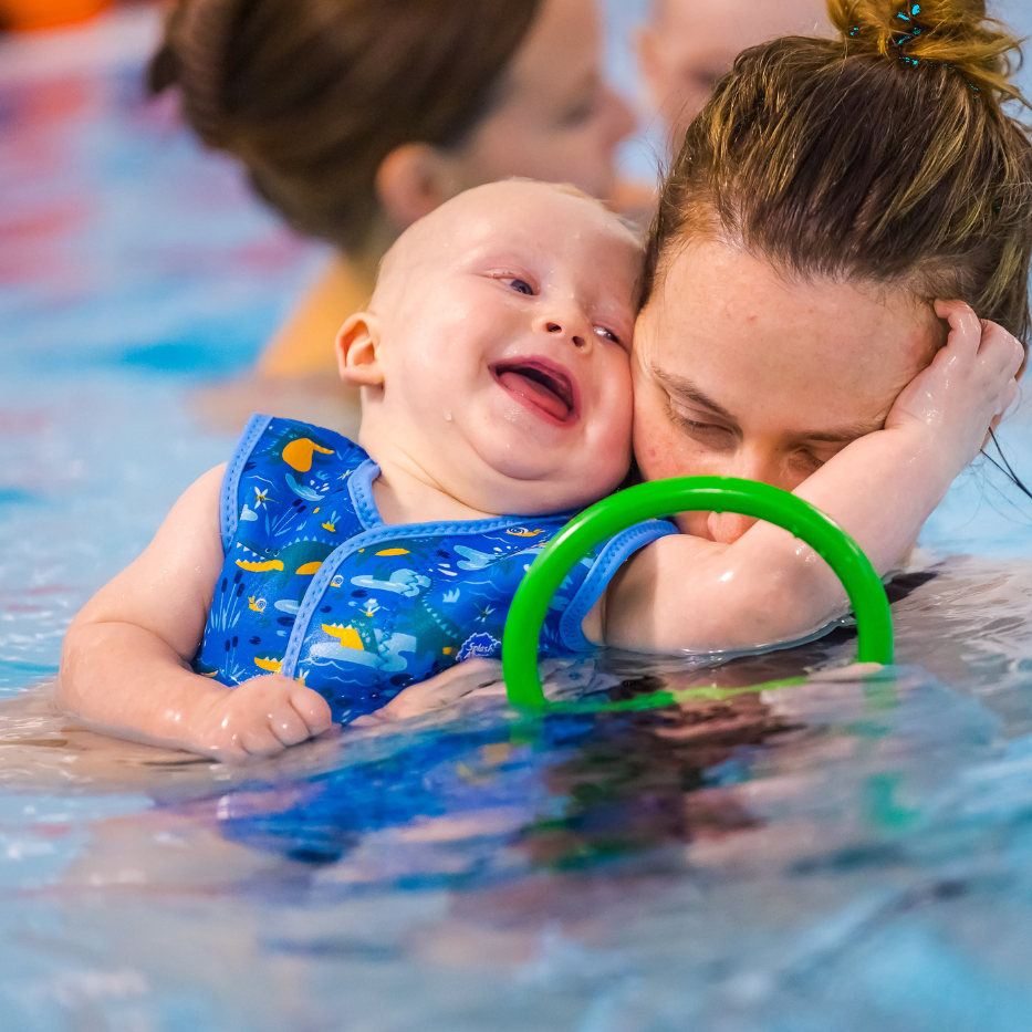 A baby smiles and cuddles their mum in the swimming pool